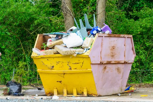 A large, yellow and pink industrial waste skip situated outdoors on a gravel surface, filled with various types of rubbish including plastic household items such as a laundry basket, a washing-up bowl, and plastic containers, along with some fabric and cardboard. The skip is positioned near a backdrop of dense green trees and bushes, suggesting a semi-rural or suburban environment. The sky appears overcast, providing diffuse lighting that highlights the textures of the accumulated waste and the weathered finish of the skip. Surrounding the skip, there are small black plastic bags and scattered debris on the ground, consistent with waste collection or removal activities, typical of commercial waste management services in the Euston area. This scene captures the tangible need for professional rubbish removal by companies like Commercial Waste Removal Euston, who handle such bulk waste clearance efficiently and responsibly, ensuring the environment is kept tidy and free from unmanaged rubbish buildup.