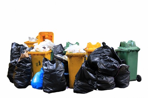 A collection of large wheeled rubbish bins and black trash bags placed outdoors on a paved surface, with some bins being yellow and others green, and the black bags appearing full and tied. The yellow bins and green bin are positioned side by side, with the black bags stacked and leaning against them, containing waste materials that are partially visible. The scene appears to be in an urban or commercial area, indicating waste collection or removal activity typical for a rubbish removal service in Euston, suggesting a focus on general waste disposal for business or residential premises. The background is plain and white, emphasizing the bins and bags as the primary objects in the image, with natural daylight illuminating the scene, highlighting the textures and colors of the waste containers and material.