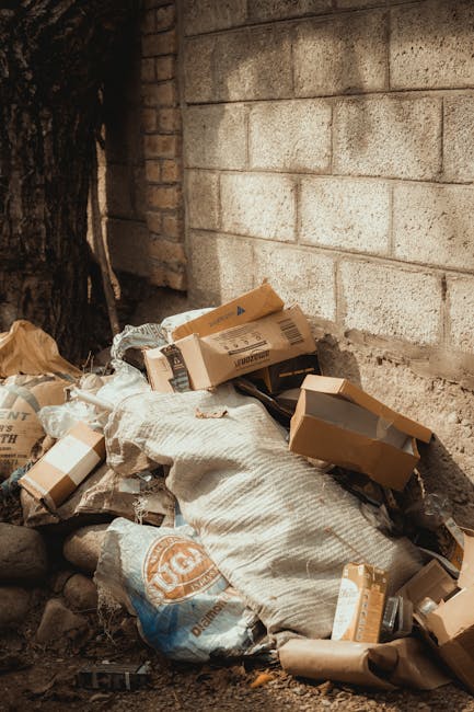 A small, makeshift cart with a metal frame and two large wheels is loaded with various discarded household items and black garbage bags. The cart contains large, box-like objects that appear to be unu