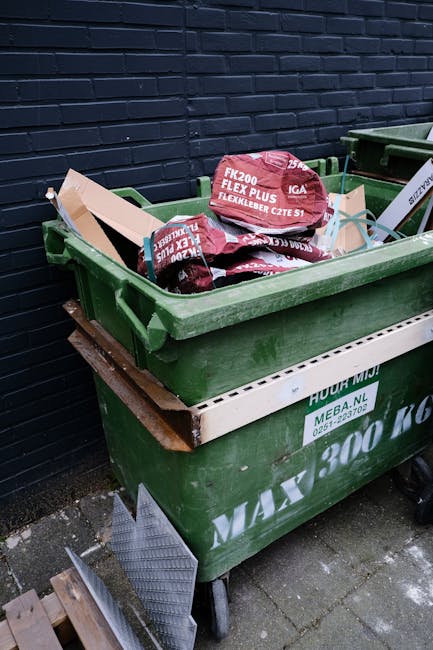 A large green wheeled rubbish bin filled with red and black bagged materials, positioned outdoors against a dark brick wall. The bin contains several plastic bags labeled 'FK200 FLEXY PLUS', with some
