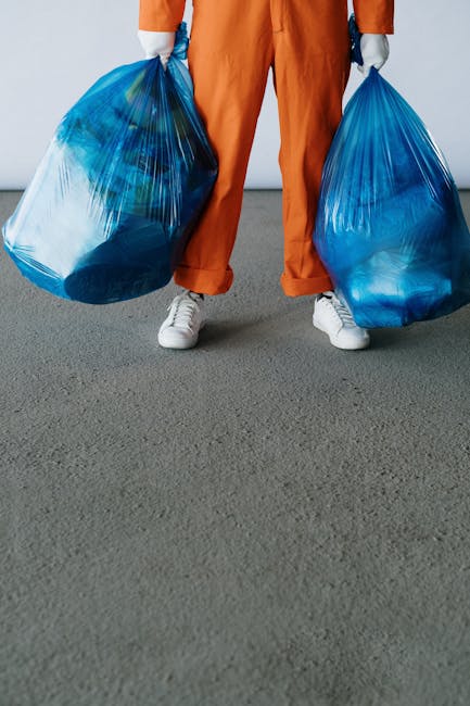 A large green wheeled rubbish bin filled with red and black bagged materials, positioned outdoors against a dark brick wall. The bin contains several plastic bags labeled 'FK200 FLEXY PLUS', with some