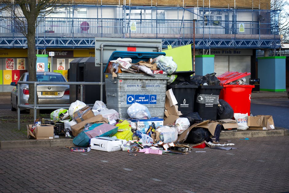 A large green waste bin filled to overflowing with various discarded cardboard boxes of different sizes and shapes, some flattened and some still assembled, along with a few wooden pallets leaning aga